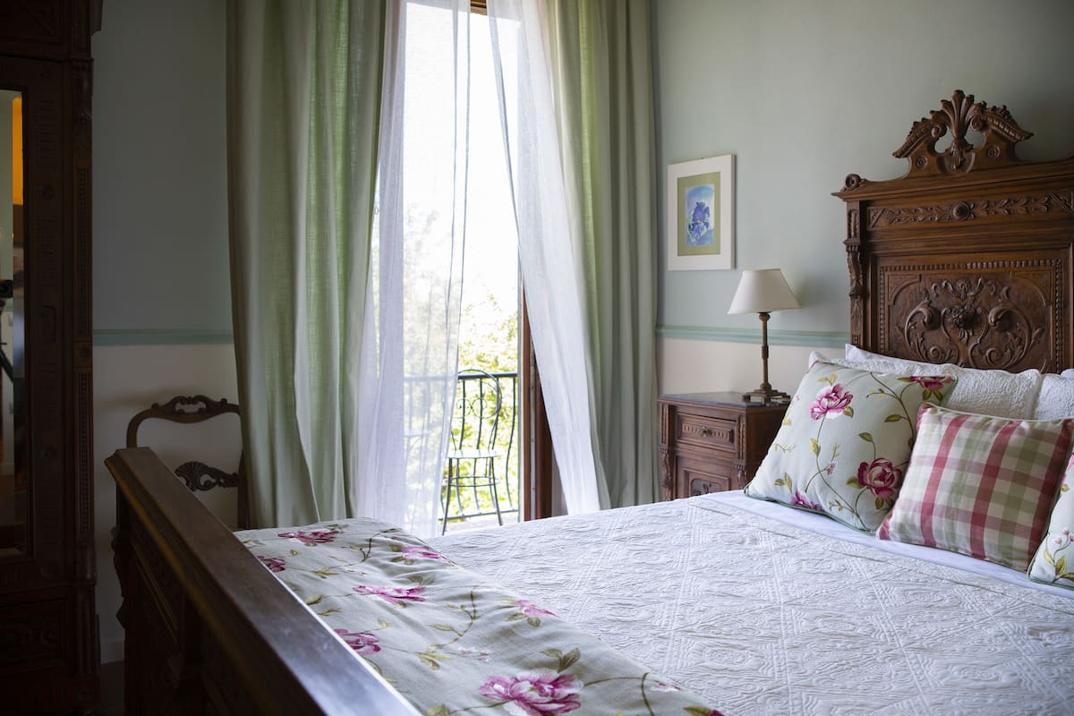 Ein Schlafzimmer mit einem hölzernen Bett, floralem Bettzeug, einem Nachttisch, einer Lampe und einem Fenster mit Vorhängen.