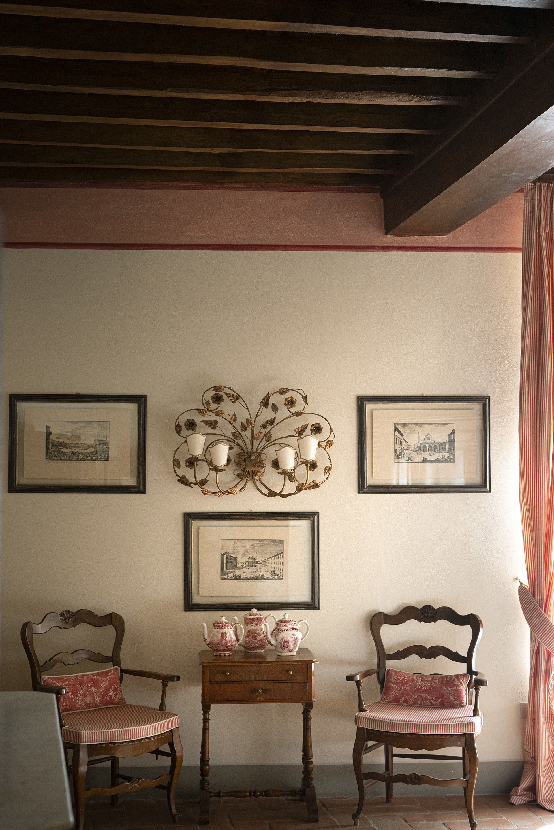 Een kamer met twee houten stoelen, een kleine tafel met een theepot en kopjes, en lijstjes met foto's aan de muur.