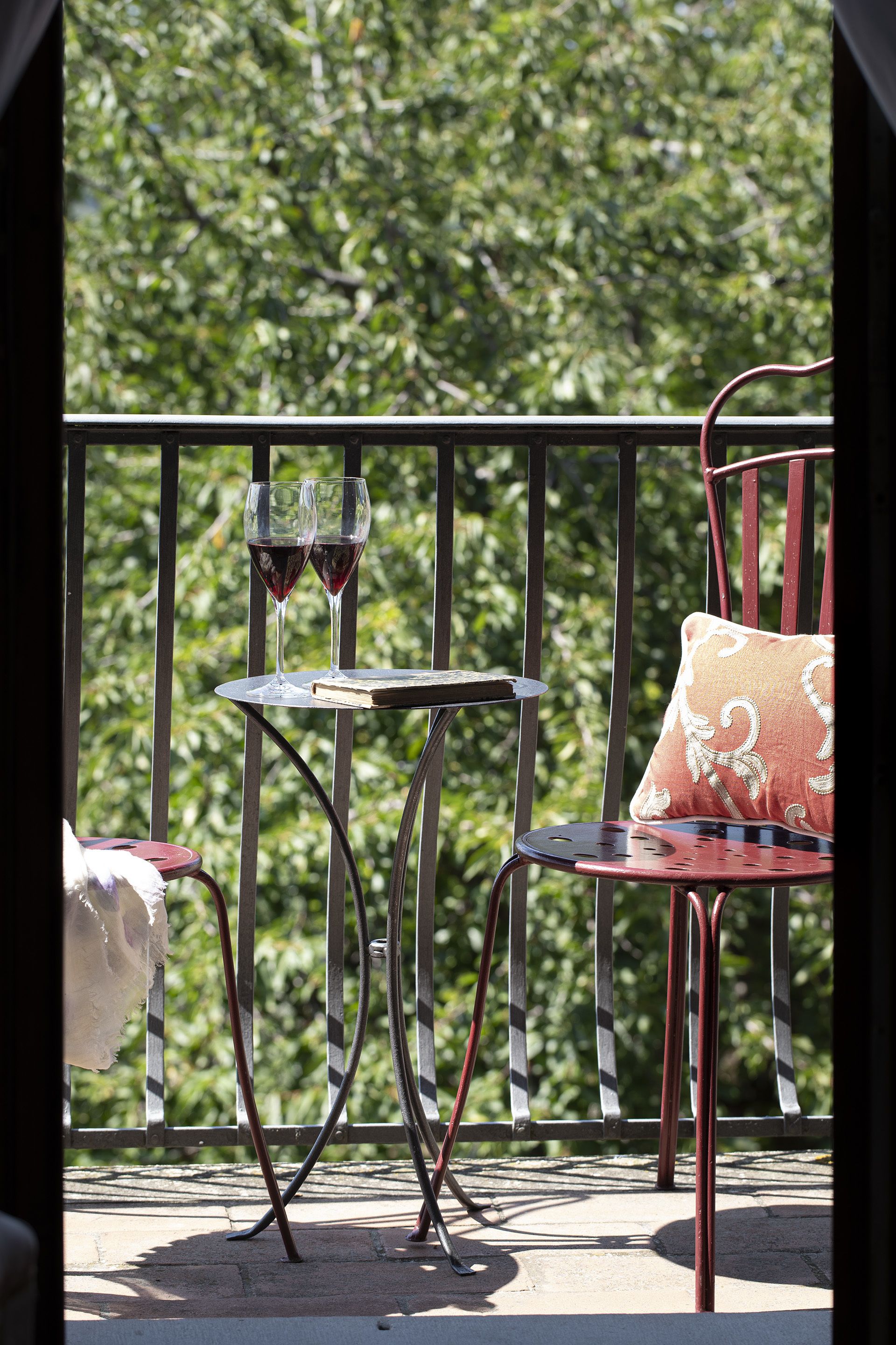 Un balcon avec une chaise en métal et une petite table, deux verres de vin rouge et un livre.