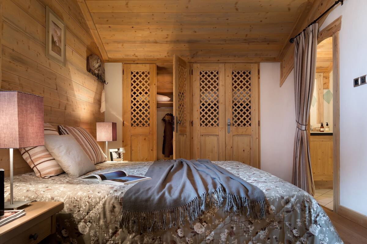 Ein gemütliches Schlafzimmer mit Holzbalkendecke, einem Bett mit floralem Bettzeug, einem Nachttisch mit Lampe und einem Kleiderschrank mit Gitterstruktur.