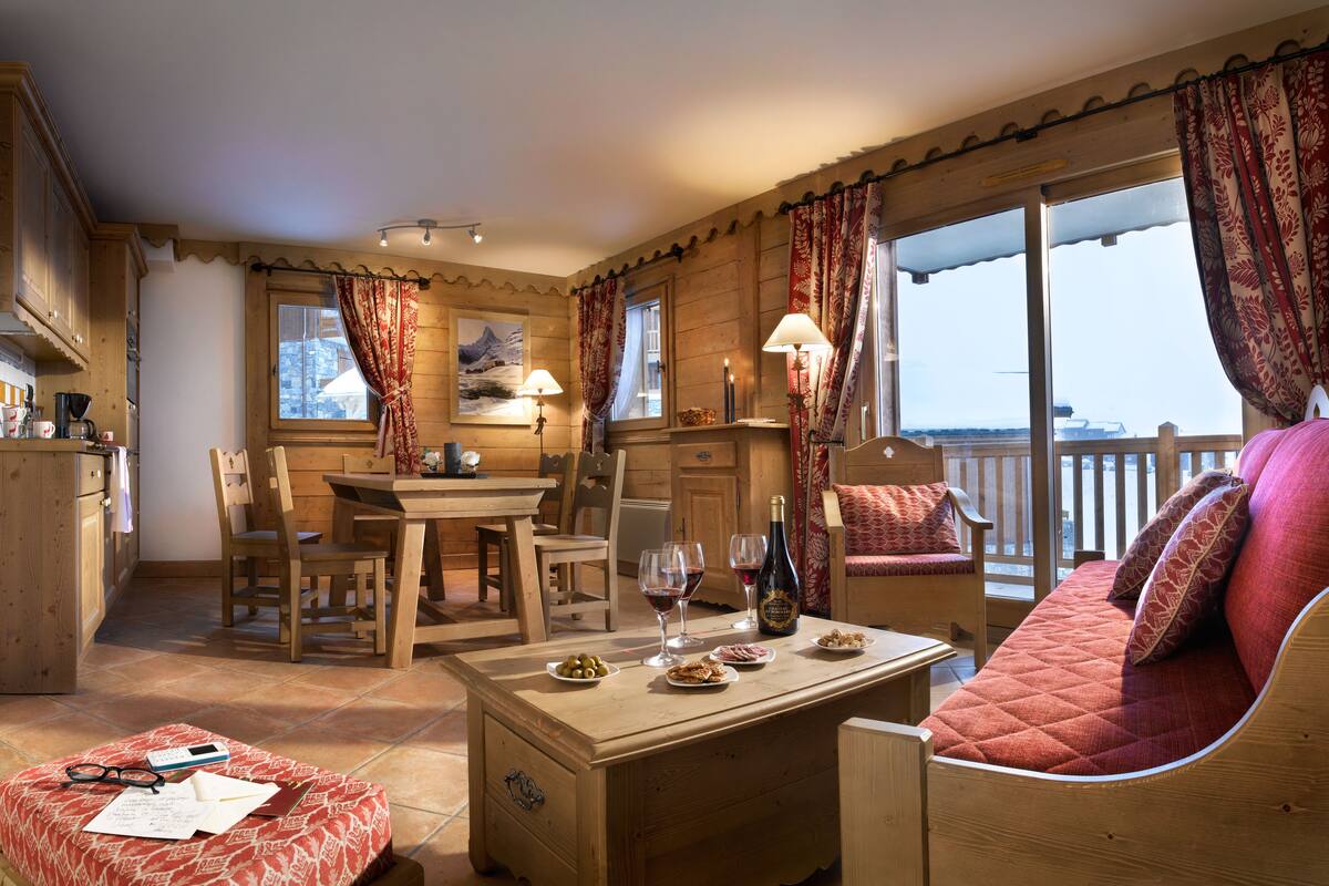 Ein gemütliches Zimmer mit Holzmöbeln, einem Esstisch und einer Couch. Zwei Fenster mit Vorhängen, Blick ins Freie, sowie ein kleiner Tisch mit Speisen und Getränken.