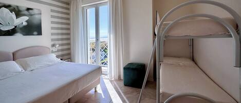 A hotel room with a bed, a bunk bed, a view of the sea, a window with curtains, and a ceiling light.