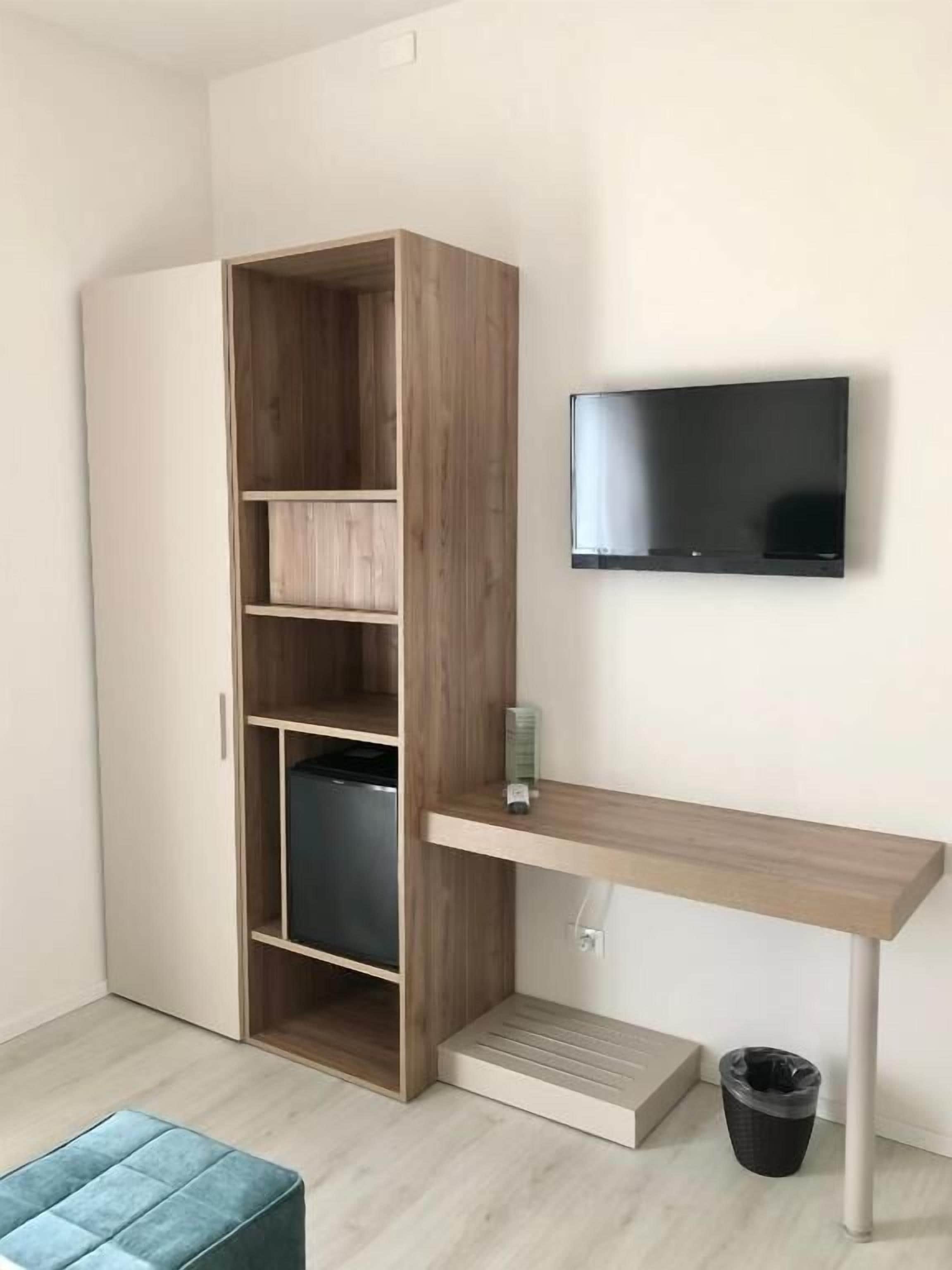 A modern room with a wooden wardrobe, a desk with a flat-screen TV, a trash bin, and a blue ottoman.