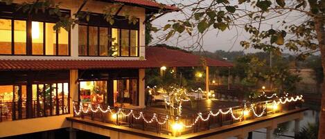 A two-story building with a red-tiled roof, a dock extending into a body of water, and string lights along the dock.
