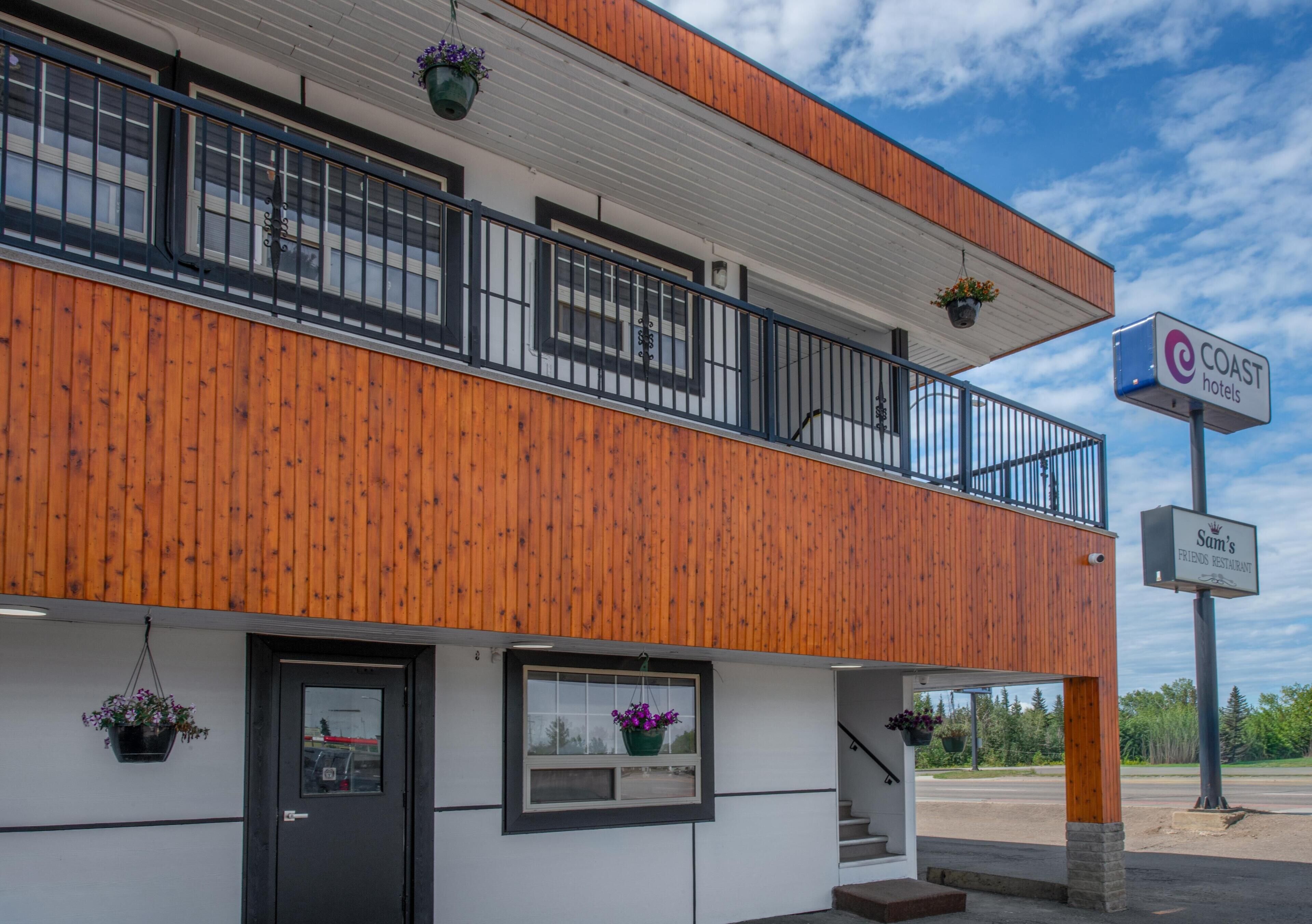 Un edificio a due piani con facciata in legno, un balcone con ringhiere nere e un cartello con la scritta 'COAST hotels' e 'Sam's Friends Restaurant'.