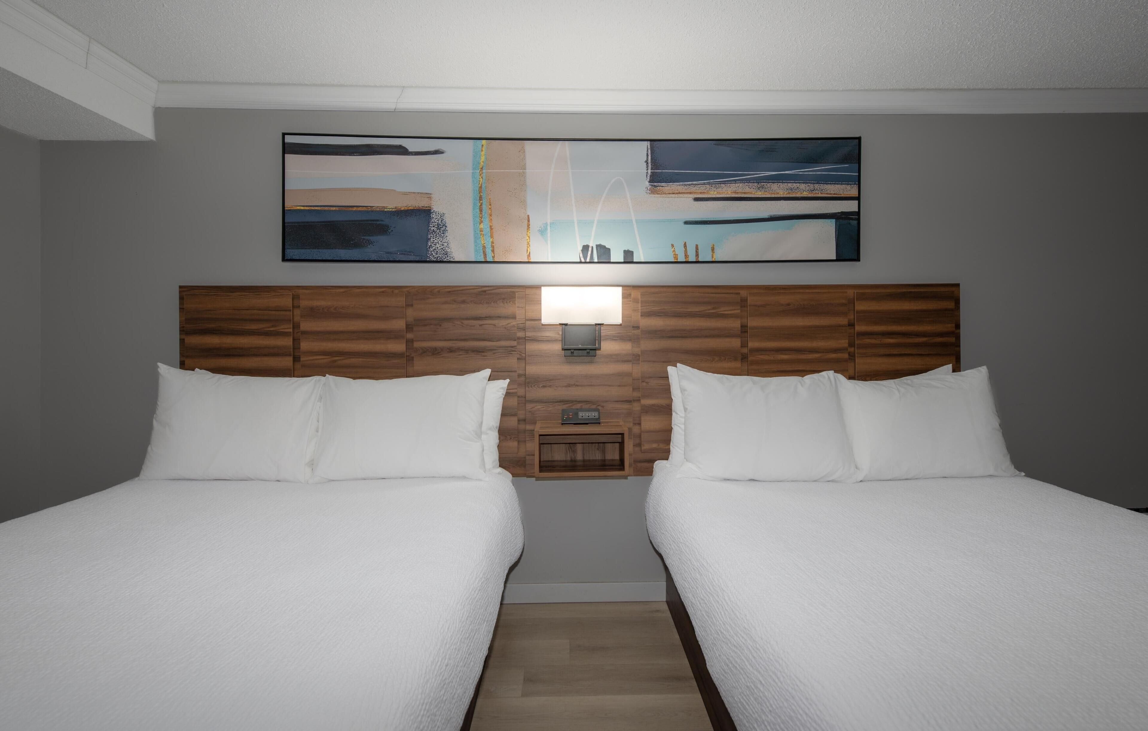 A hotel room with two beds, a wooden headboard, a wall-mounted artwork, and a nightstand with a lamp.