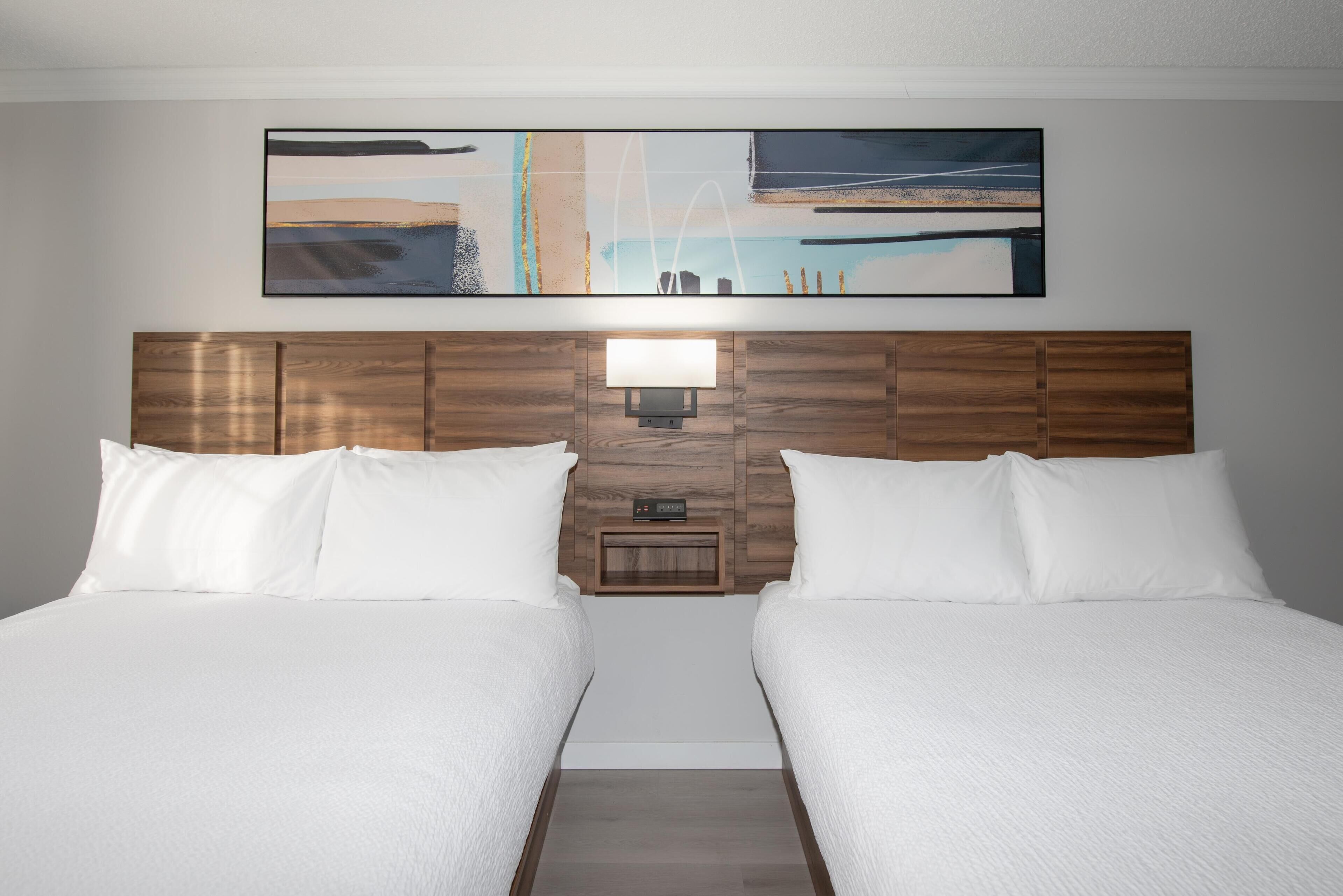 Een hotelkamer met twee bedden, een houten hoofdeinde, een wandlamp en een groot abstract schilderij boven de bedden.