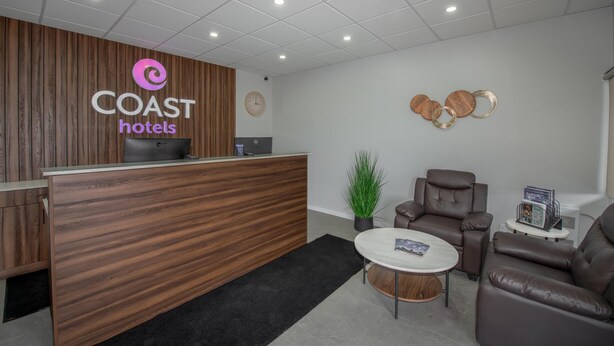A reception desk with a wooden finish, a clock, a potted plant, leather seating, and a small table with magazines.