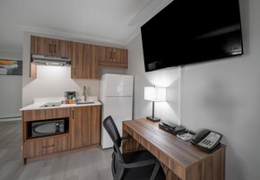 Uma cozinha compacta com micro-ondas, geladeira e cafeteira, uma mesa de escritório com luminária e telefone, e uma TV fixada na parede.