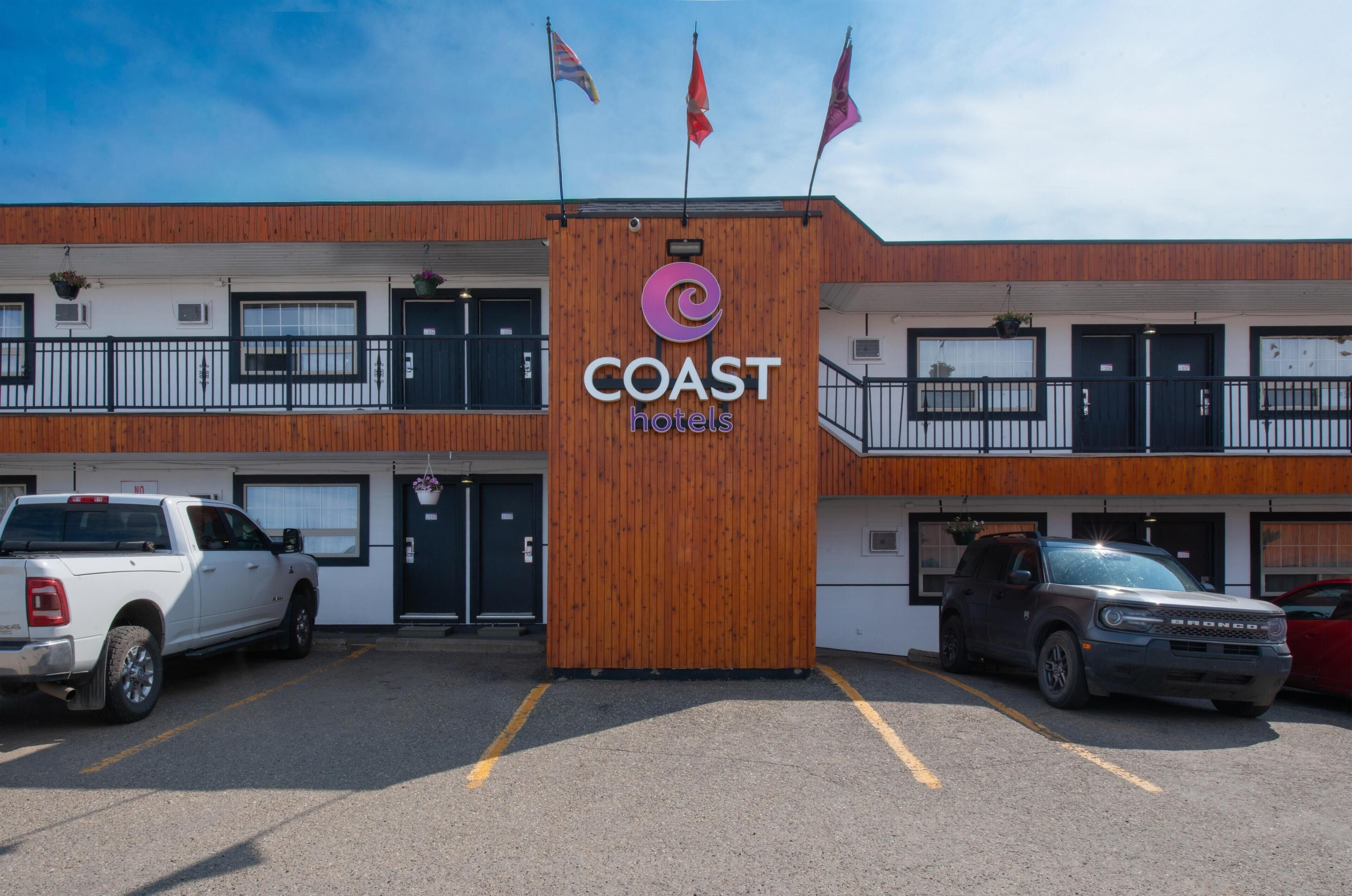 Un bâtiment à deux étages, sur lequel on peut voir une enseigne indiquant « COAST Hotels », et un parking où sont stationnés plusieurs véhicules.