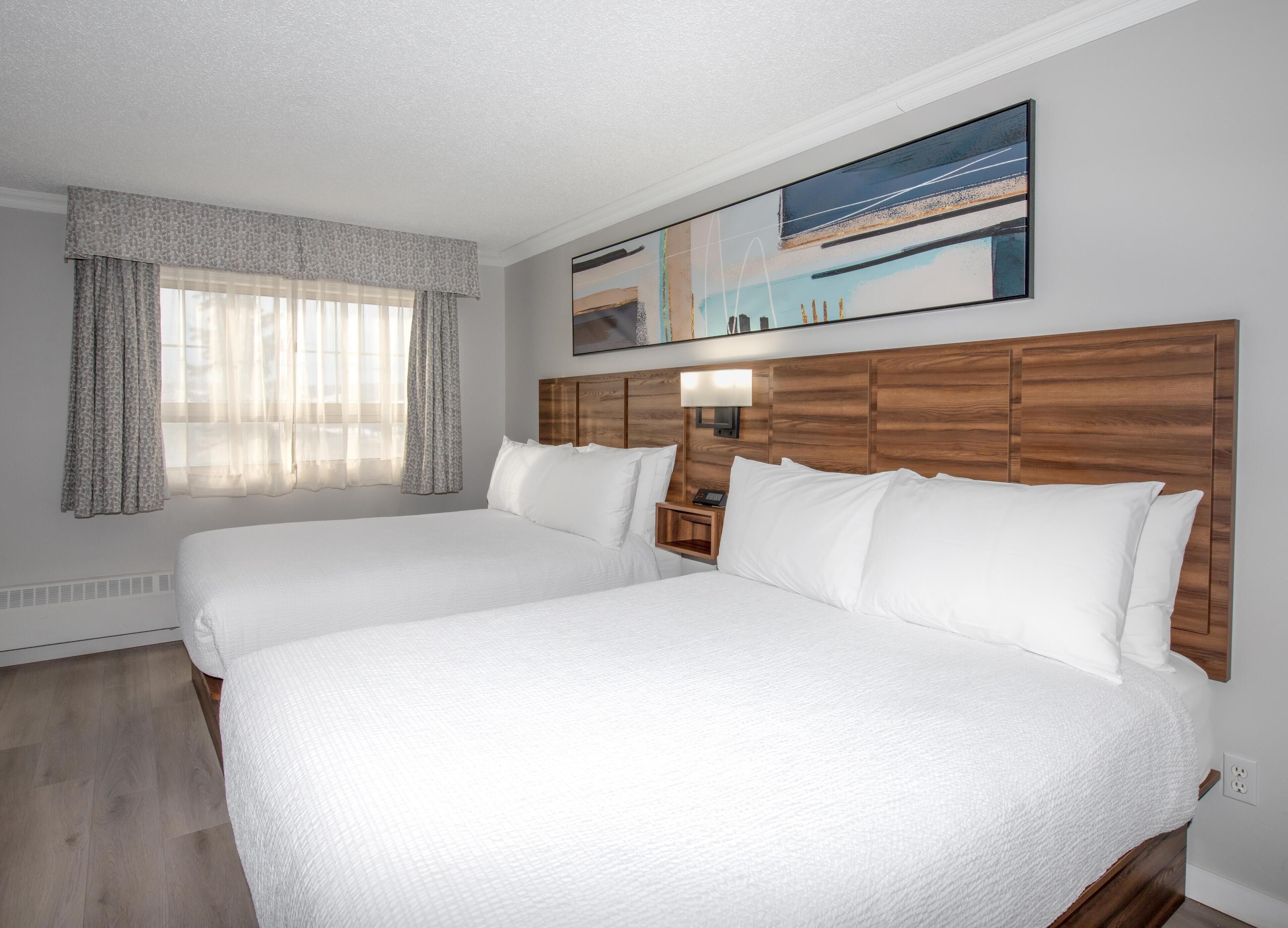 Habitación de hotel con dos camas, cabecera de madera, mesita de noche, un cuadro y una ventana con cortinas traslúcidas.