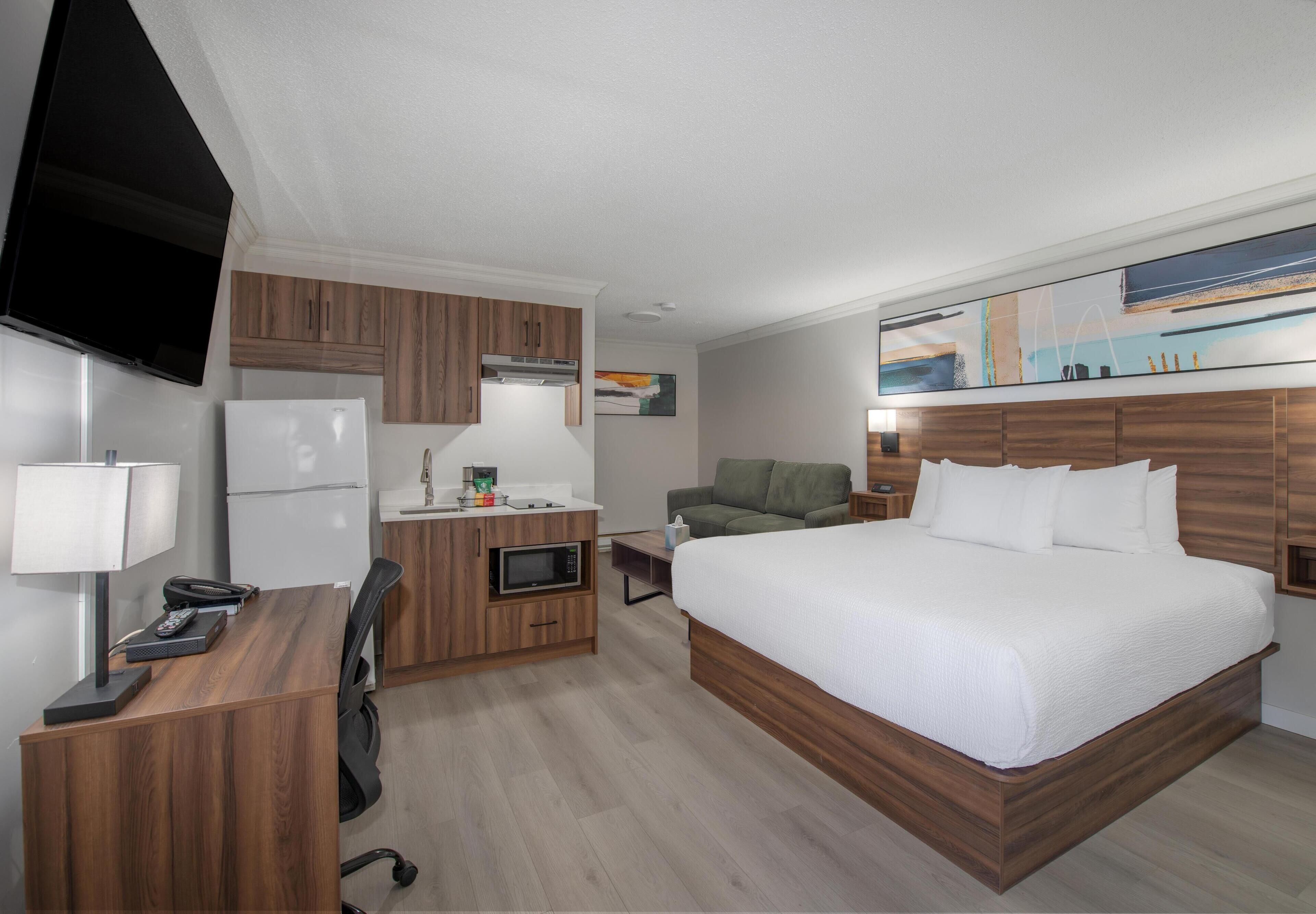 A hotel room with a bed, a desk, a kitchenette, a TV, and a painting on the wall.