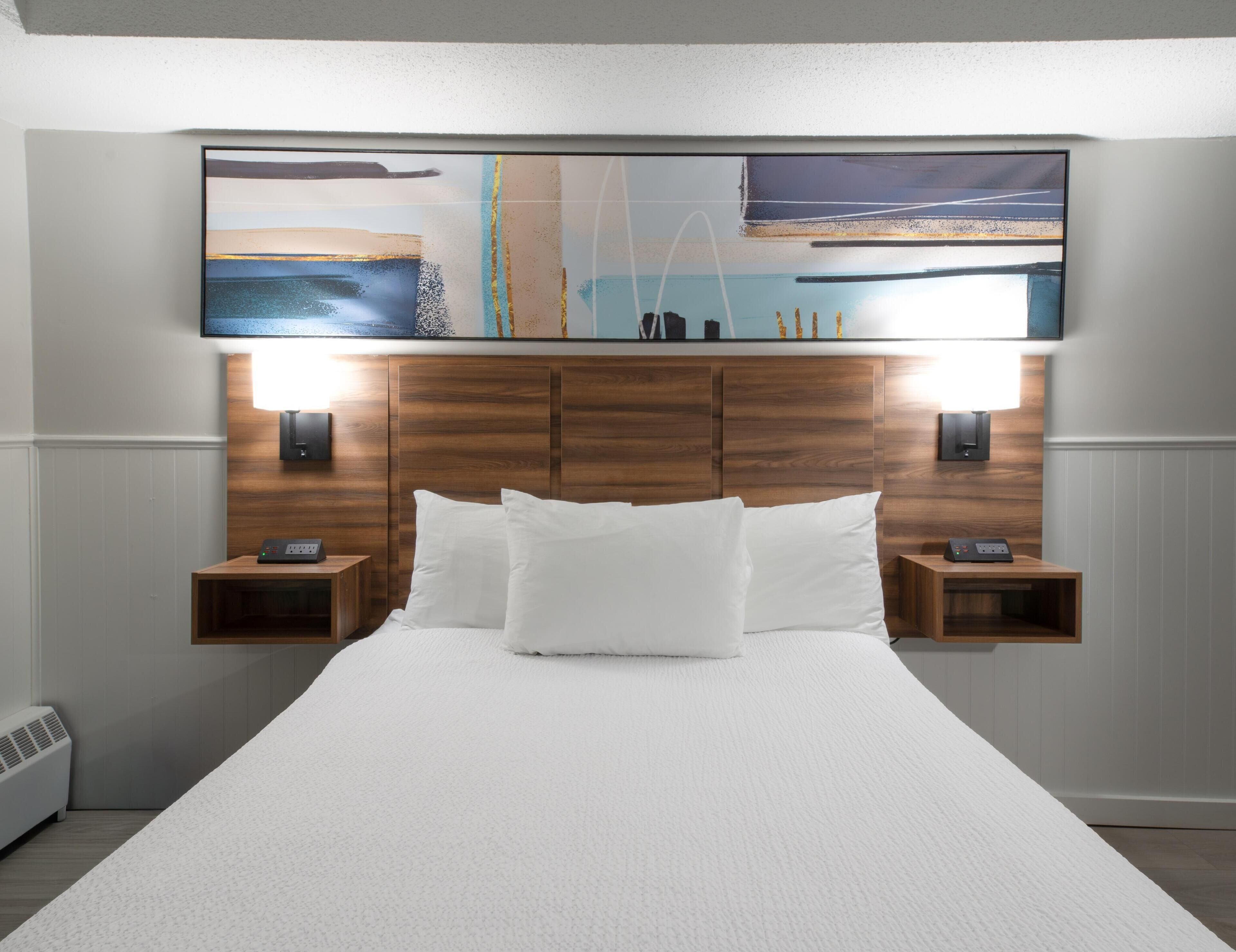 Habitación de hotel con una cama grande, cabecera de madera, dos mesitas de noche con lámparas y una obra de arte colgada en la pared sobre la cama.
