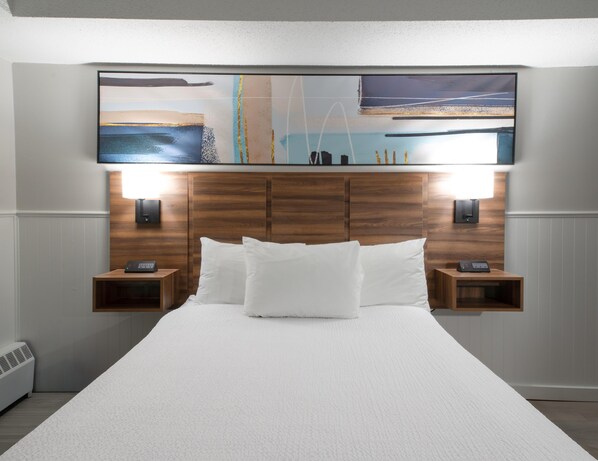 Habitación de hotel con una cama grande, cabecera de madera, dos mesitas de noche con lámparas y una obra de arte colgada en la pared sobre la cama.