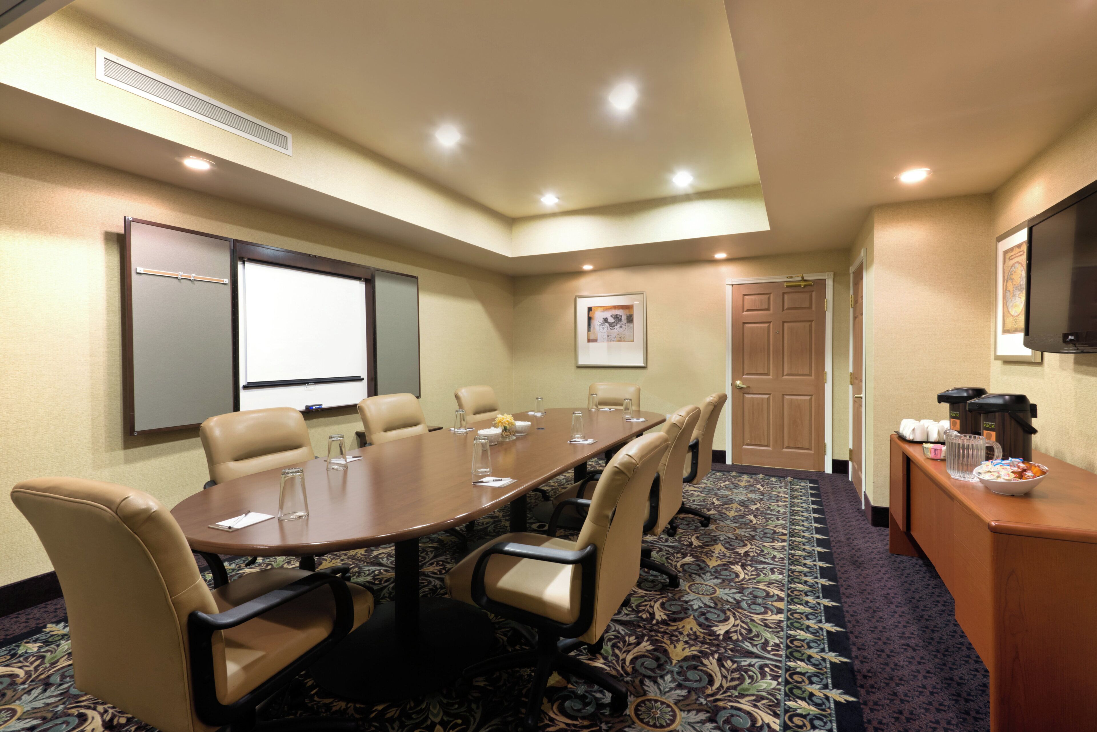 A conference room with a long table, chairs, a projector screen, a door, and a television.