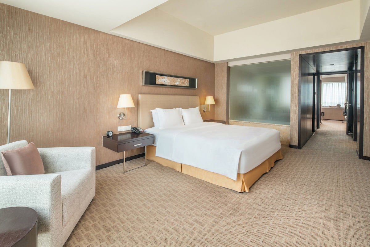 Ein Hotelzimmer mit einem großen Bett, einem Schreibtisch mit Lampe, einem Sessel und einem großen Fenster.