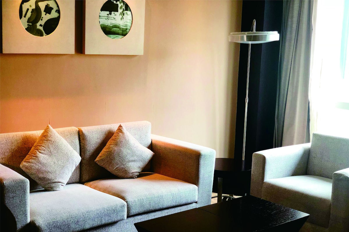 Ein modernes Wohnzimmer mit einem grauen Sofa, zwei Kissen, einem schwarzen Couchtisch und einer Stehlampe. An der Wand hängen zwei gerahmte Bilder.