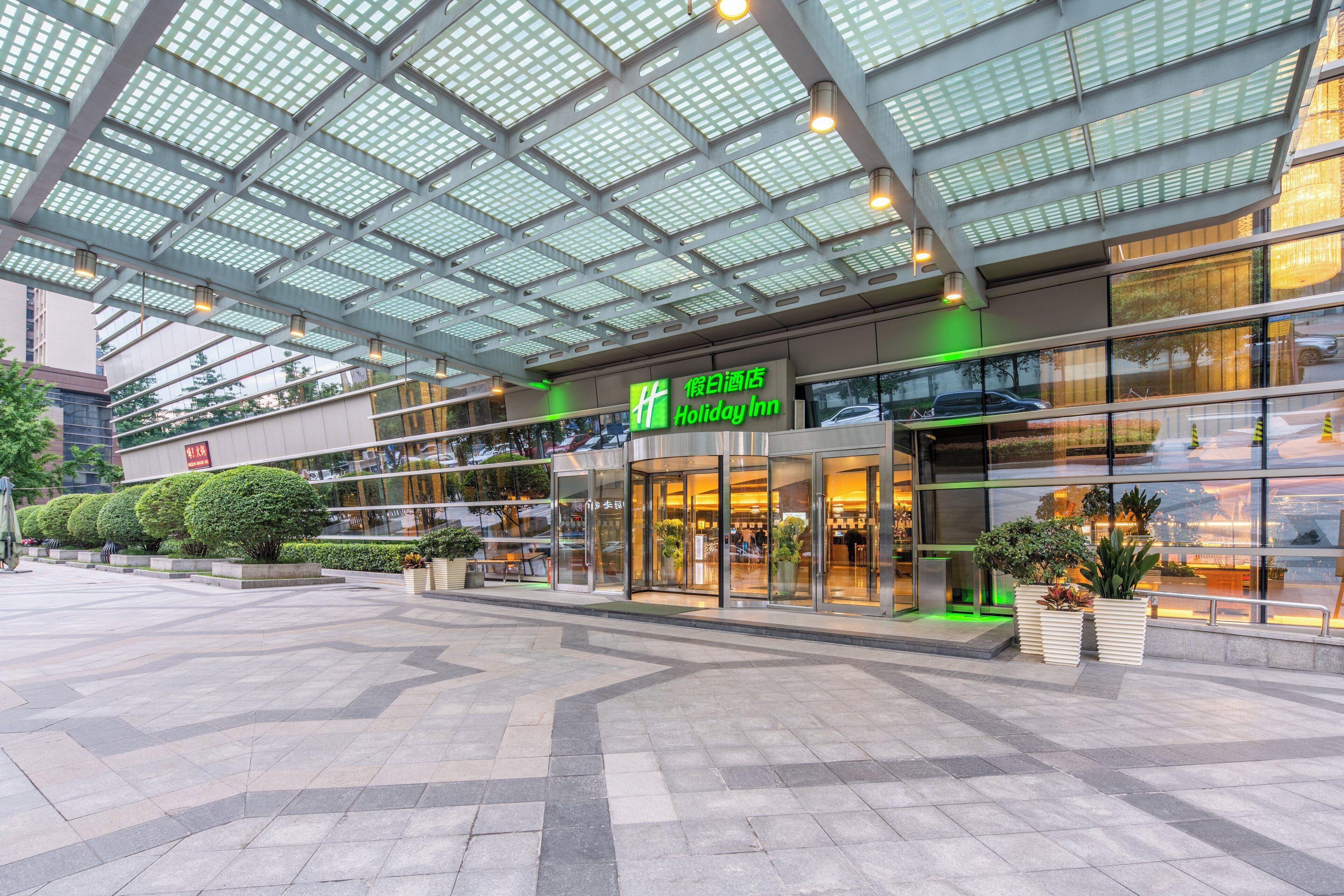 A Holiday Inn hotel with a modern glass facade, outdoor seating area, and a covered walkway.