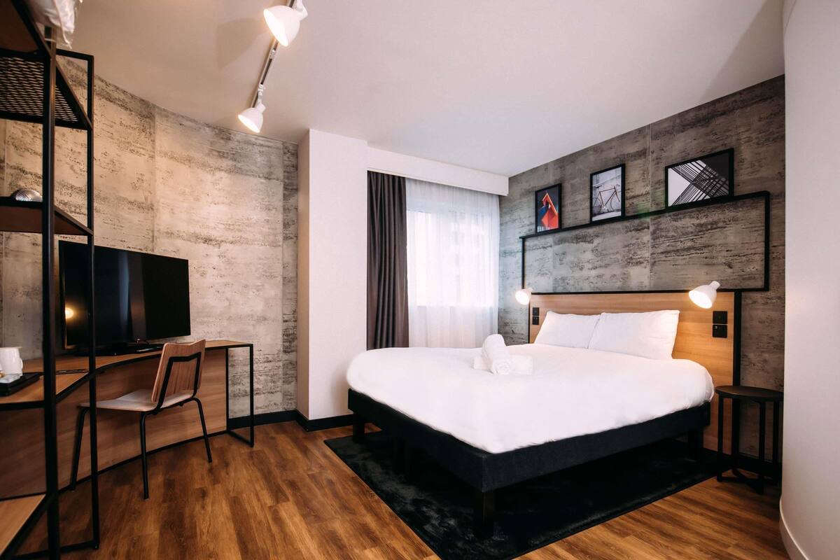 A modern hotel room with a large bed, a desk with a chair, a television, and a wooden floor.