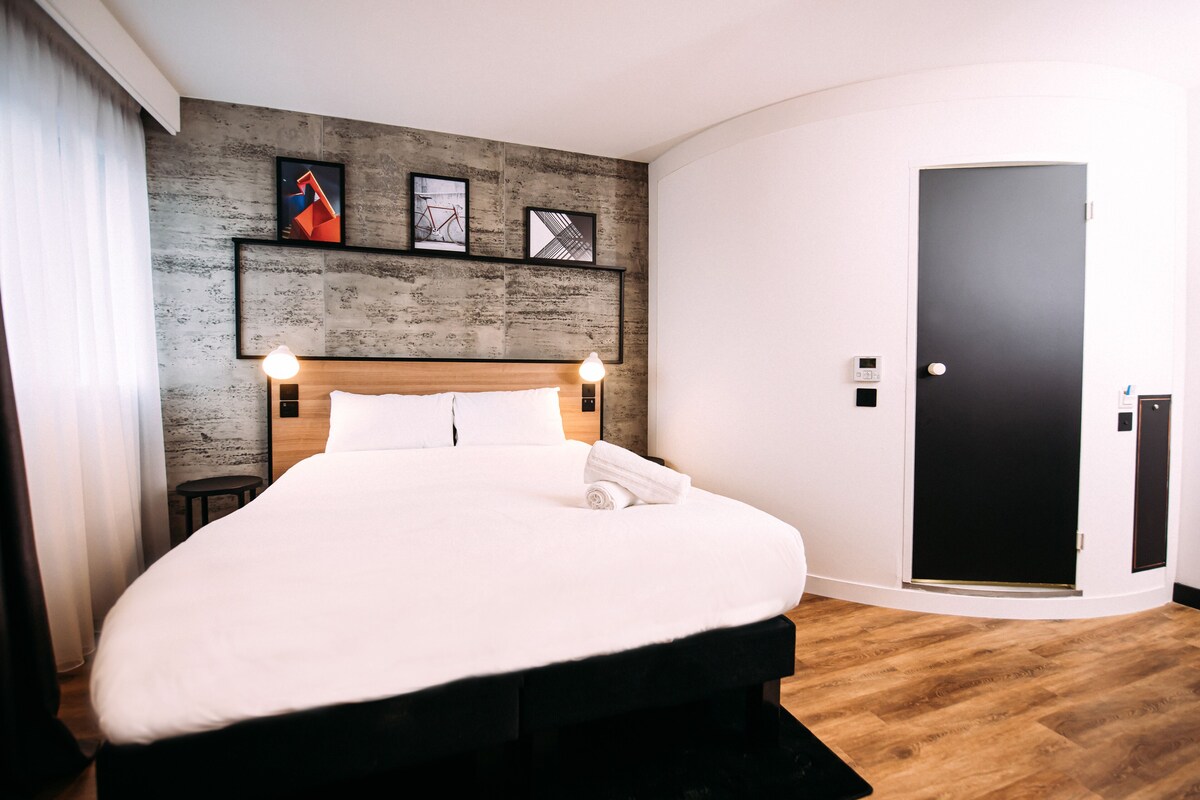 A modern hotel room with a large bed, wooden flooring, a dark door, and framed artwork on the wall.