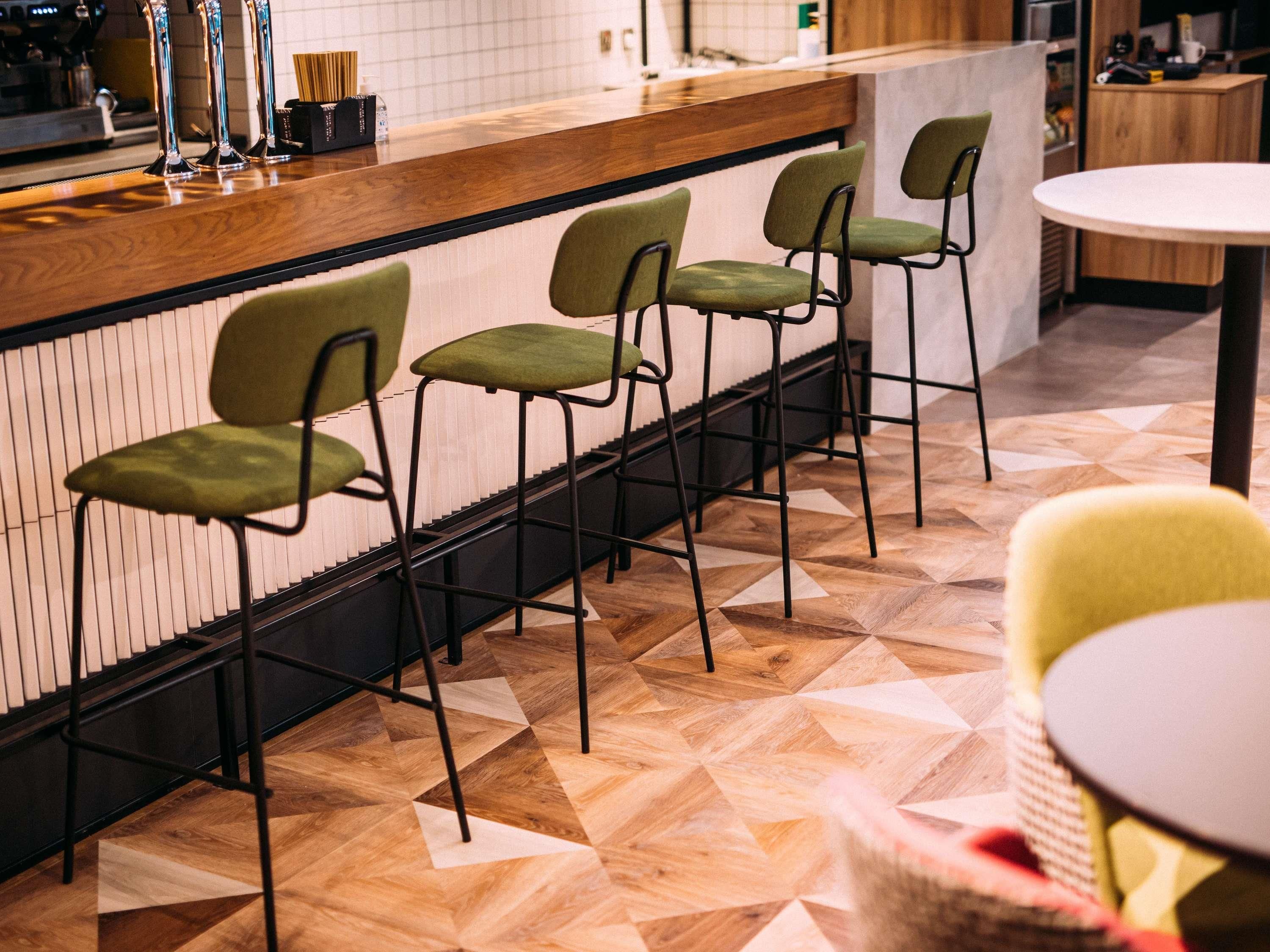 Una zona de bar con mostrador de madera, sillas con reposabrazos verdes y suelo estampado.