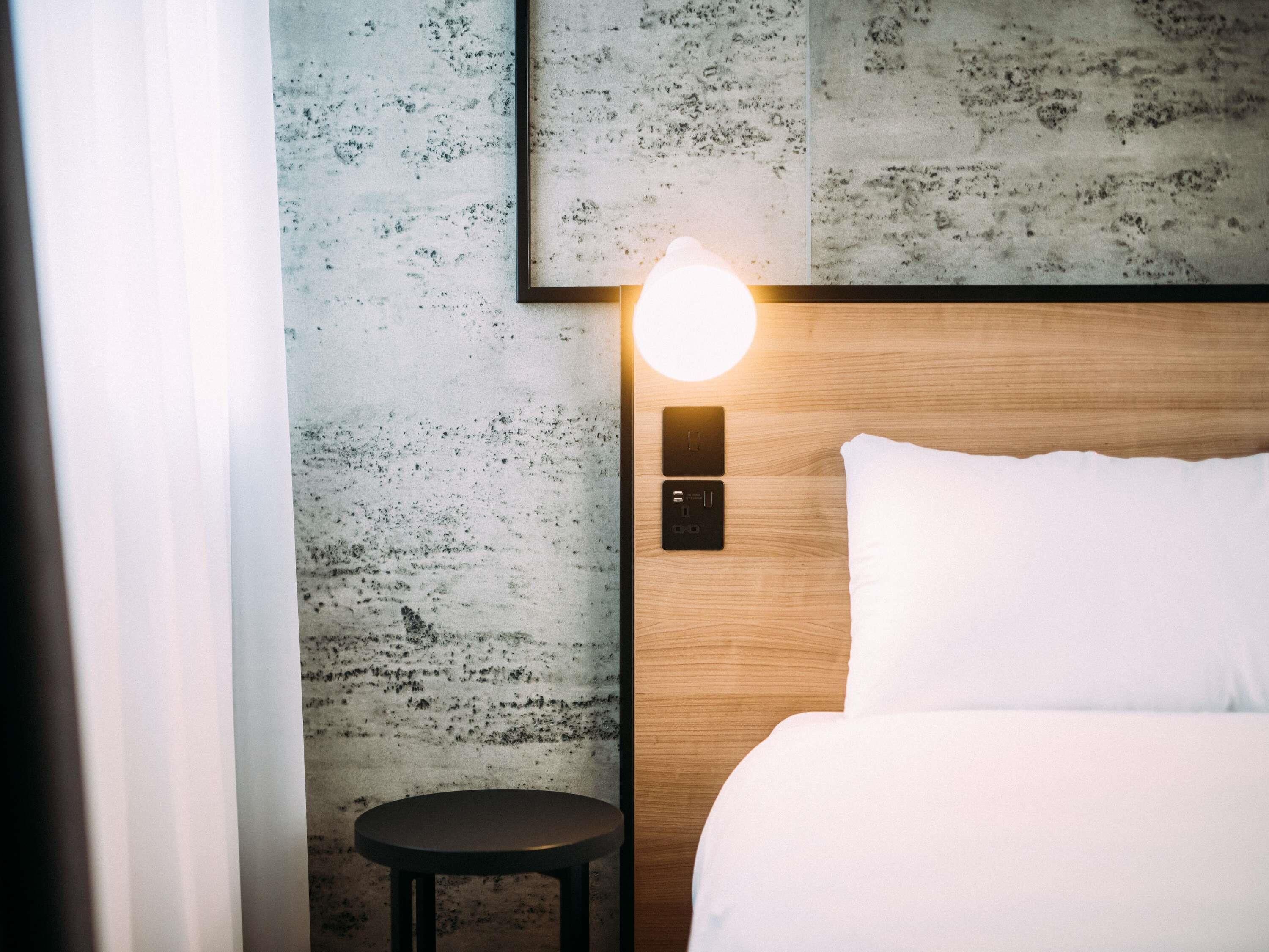 Una habitación de hotel con una cama, una mesita redonda pequeña, una lámpara de pared y un panel de control.