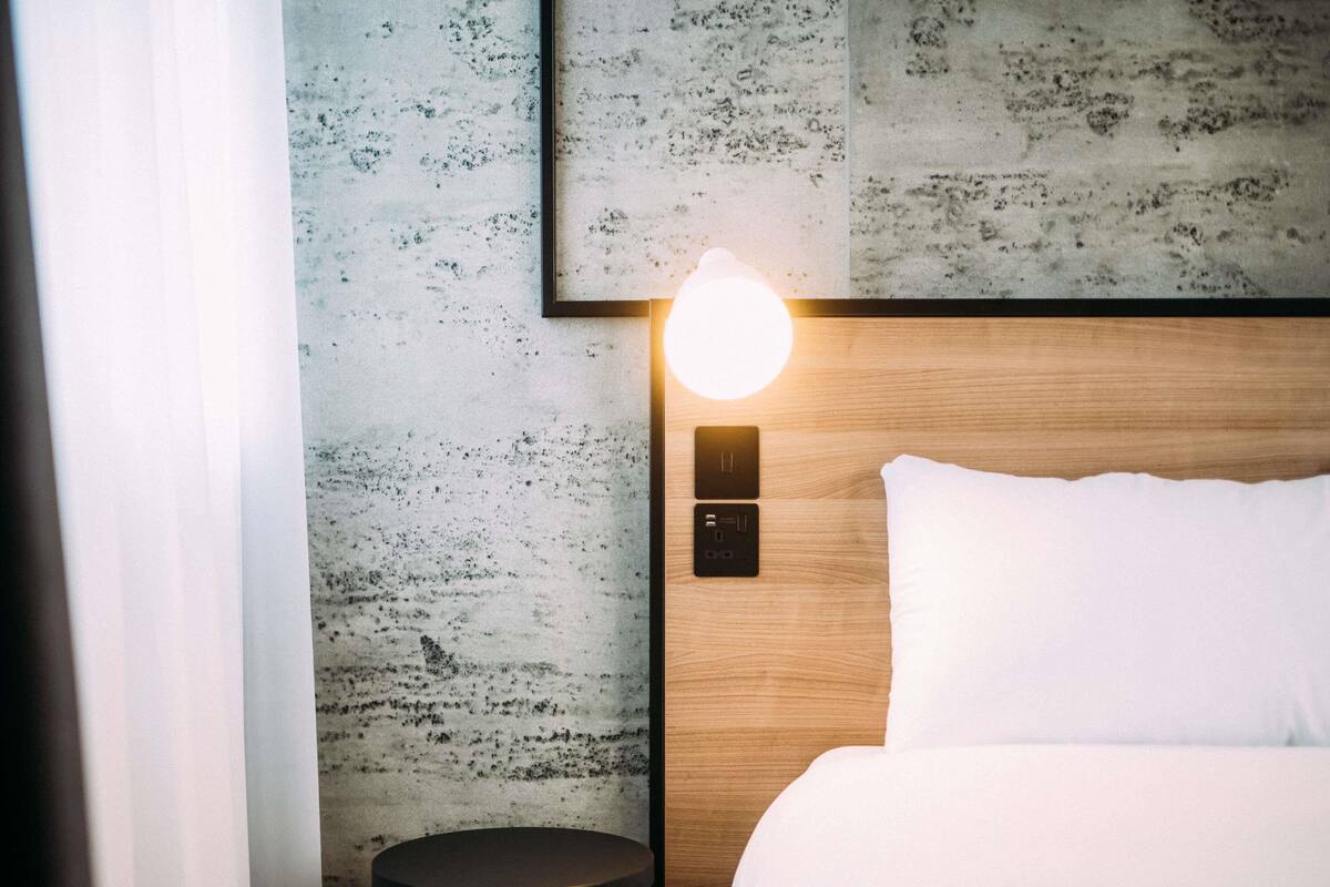 A hotel room with a bed, a small round table, a wall-mounted light, and a control panel.