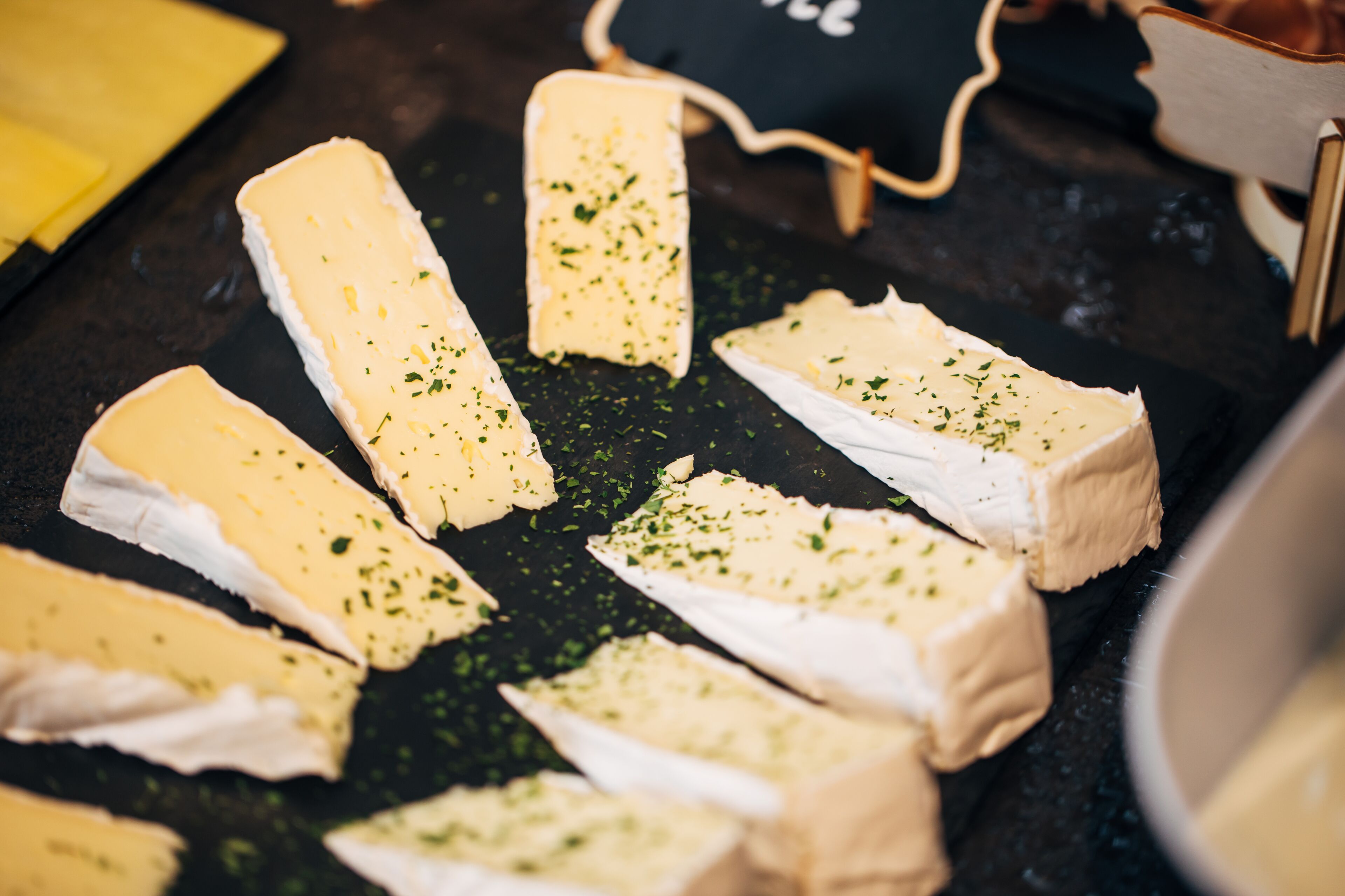 Varias porciones de queso sobre una superficie oscura, posiblemente una tabla de cortar, con algunas hierbas verdes espolvoreadas encima.