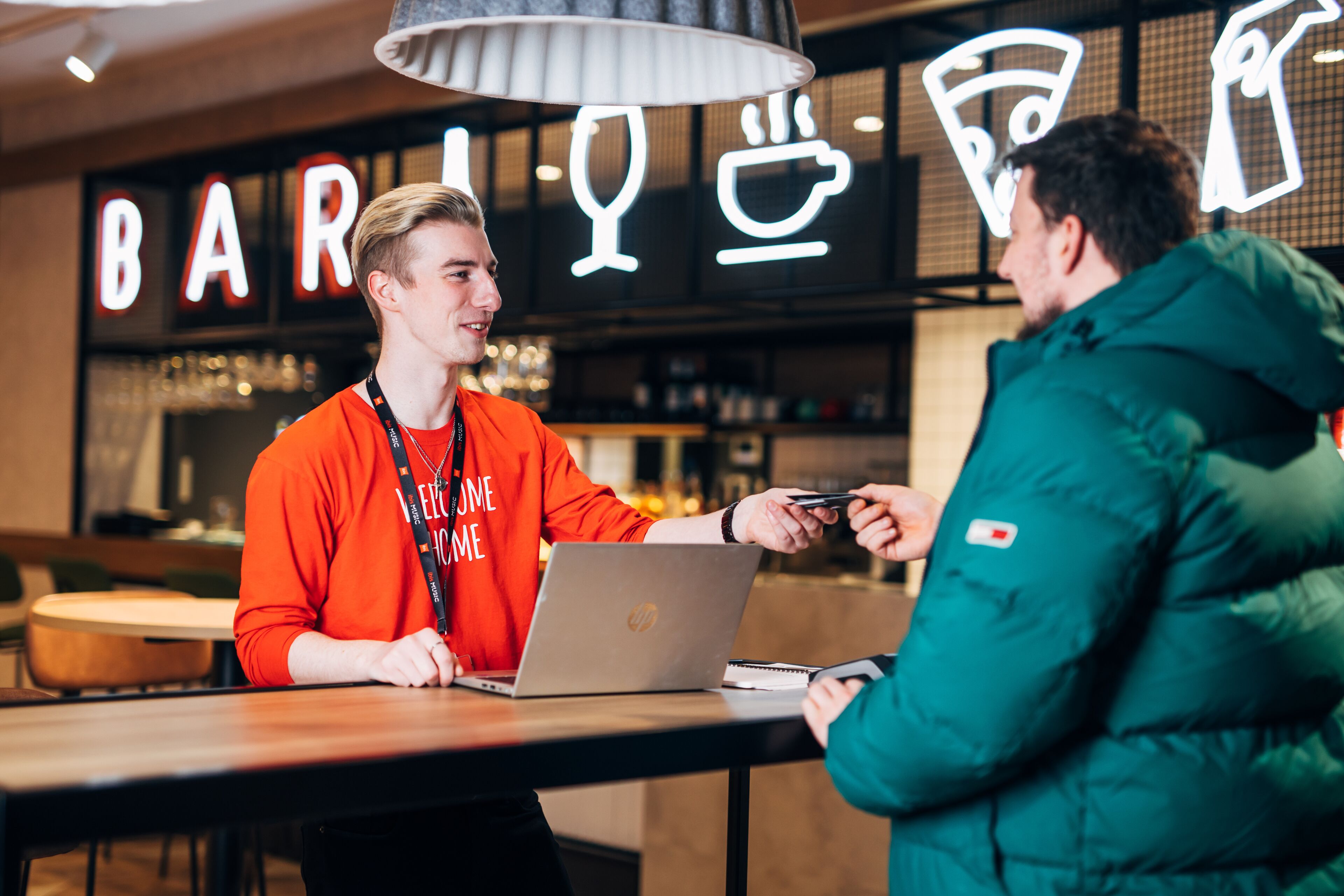 A person in an orange shirt sitting at a table with a laptop, another person in a green jacket standing beside them, and a bar sign in the background.