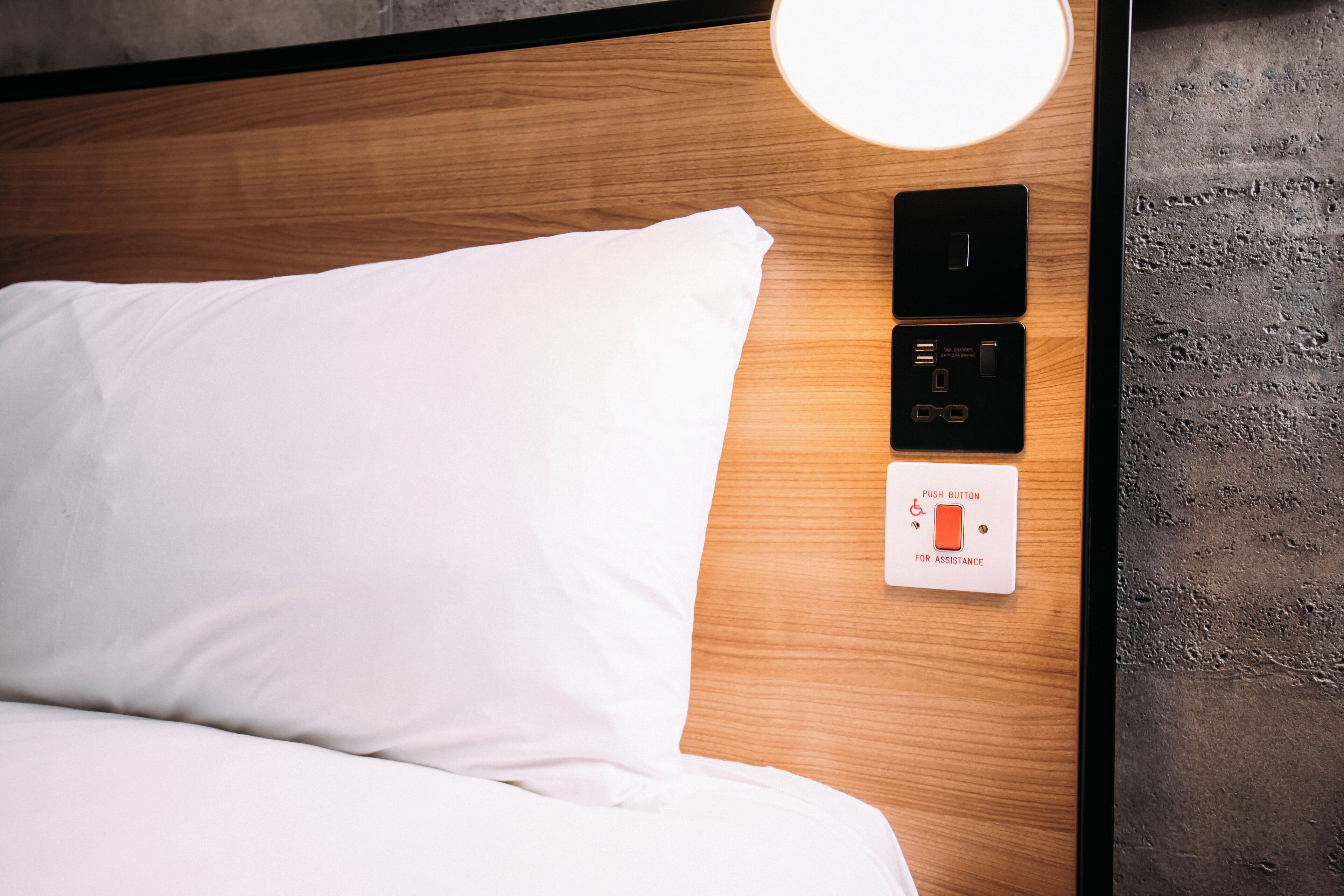 Een netjes opgemaakt bed met witte lakens, een houten hoofdbord en een bedieningspaneel aan de muur met een drukknoop voor hulp.