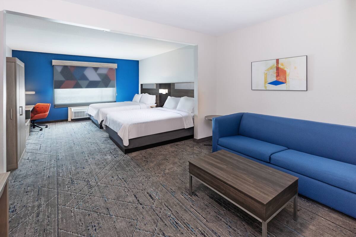 A modern hotel room with two beds, a blue sofa, a wooden coffee table, and a geometric wall art piece.