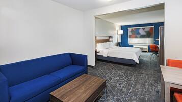 Habitación de hotel con sofá azul, una mesita de centro de madera, una cama con ropa de cama blanca, un escritorio con una silla y un ventanal con vistas geométricas.