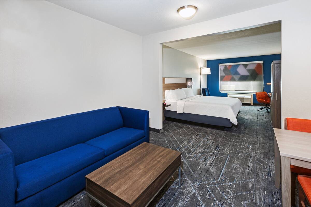 A hotel room with a blue sofa, a wooden coffee table, a bed with white linens, a desk with a chair, and a large abstract painting on the wall.
