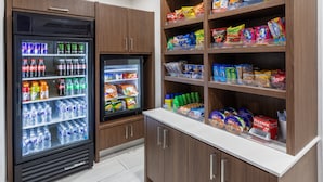 Espaço com lanches e bebidas, incluindo bebidas geladas e prateleiras repletas de petiscos e água engarrafada.
