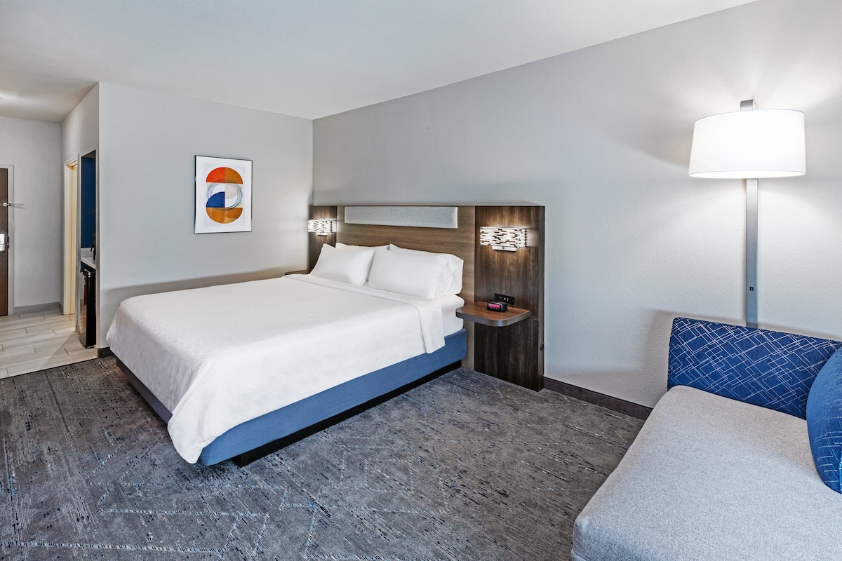 A modern hotel room with a large bed, a bedside table with a lamp, and a wall-mounted artwork.