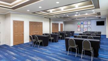 Un salón de conferencias con varias mesas y sillas, piso alfombrado y una pared decorada con motivos enmarcados.