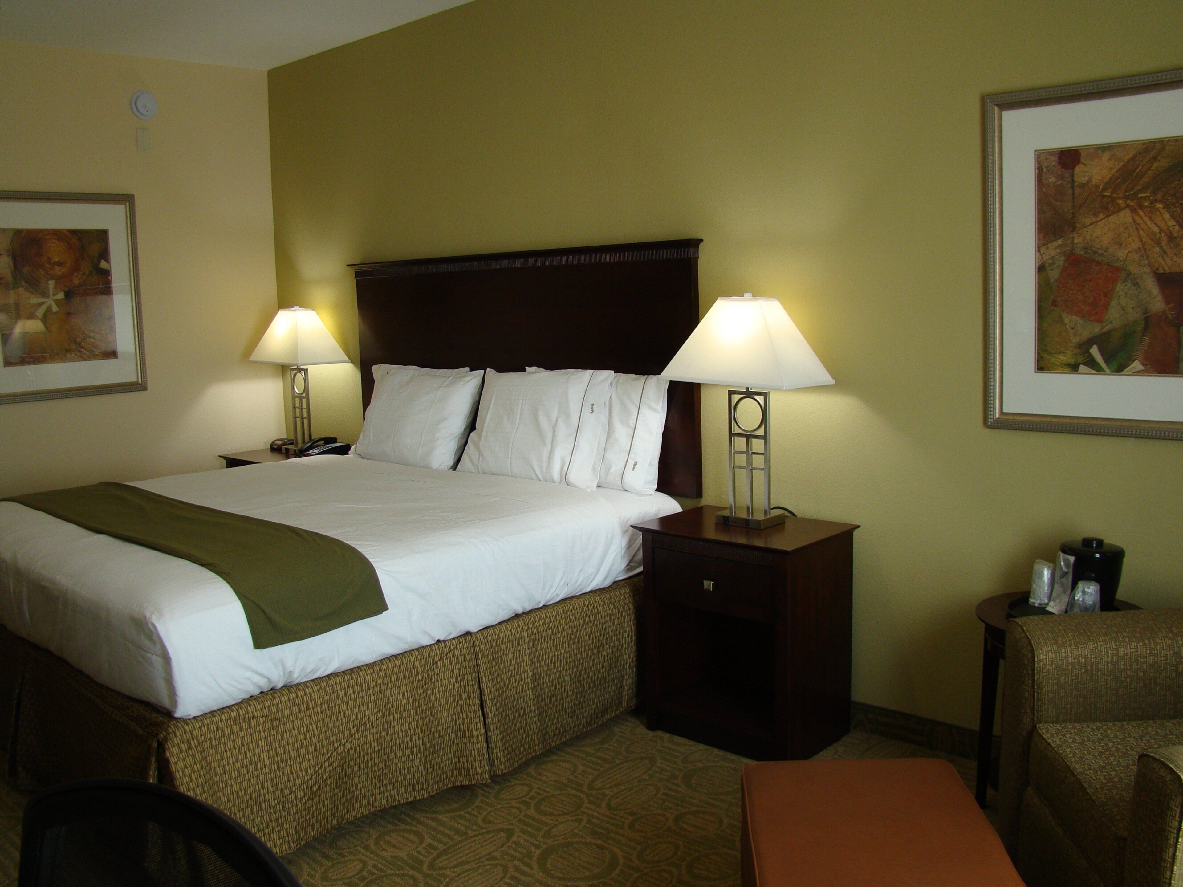 A hotel room with a large bed, two bedside lamps, a nightstand, a chair, and two framed artworks on the walls.