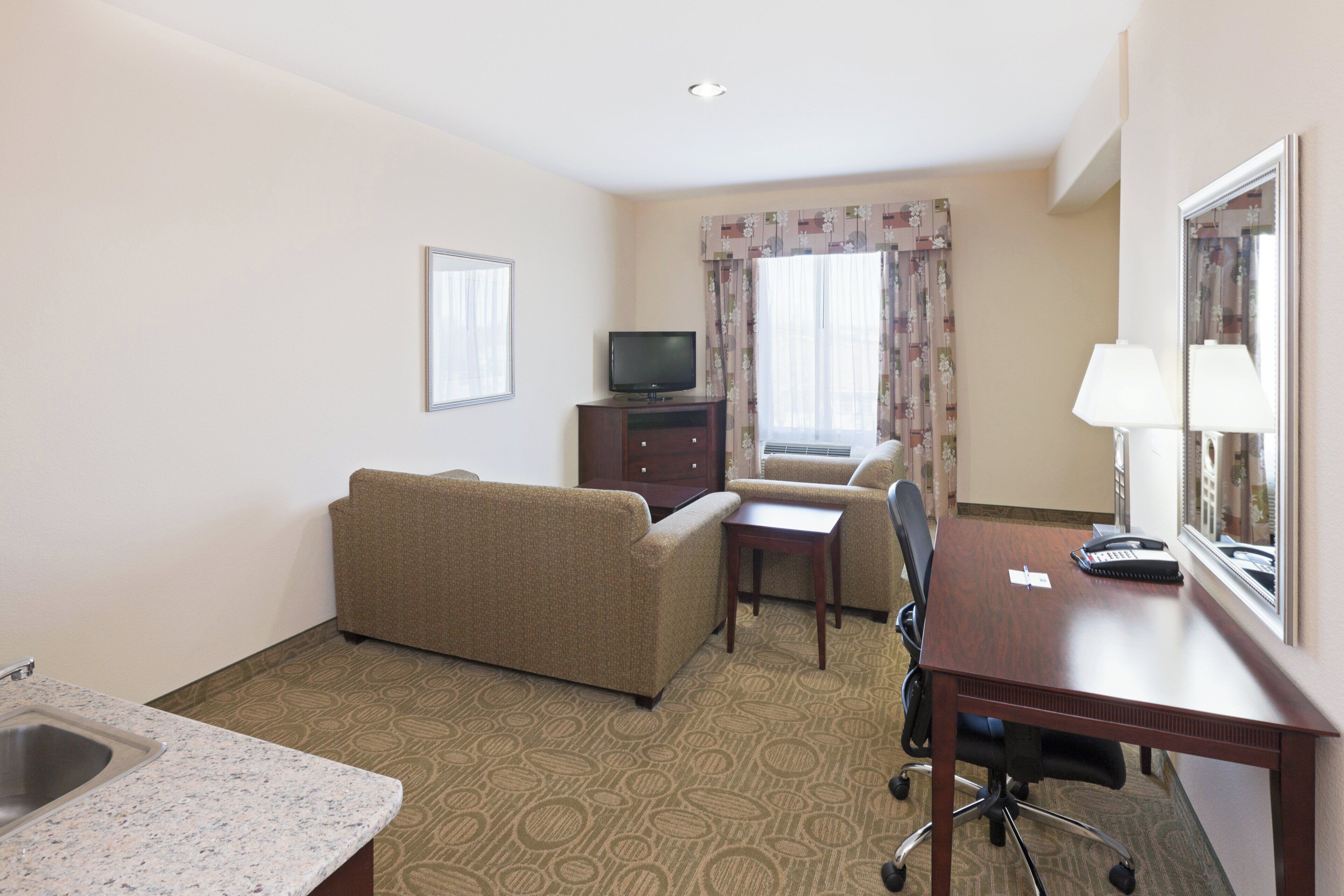 A hotel room with a sofa, a small table, a desk with a telephone, a television, a mirror, and a window with curtains.