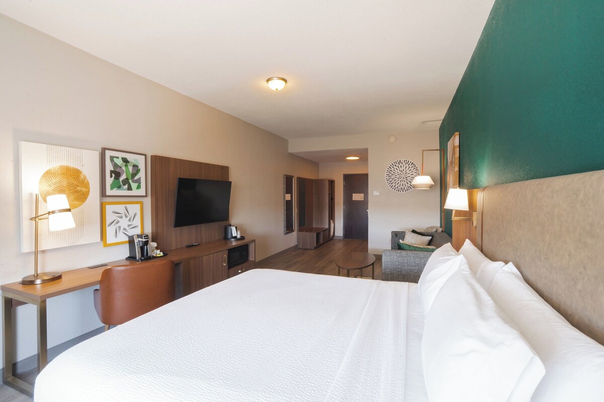 A hotel room with a large bed, a desk with a lamp, a television, and a seating area with a chair and a table.