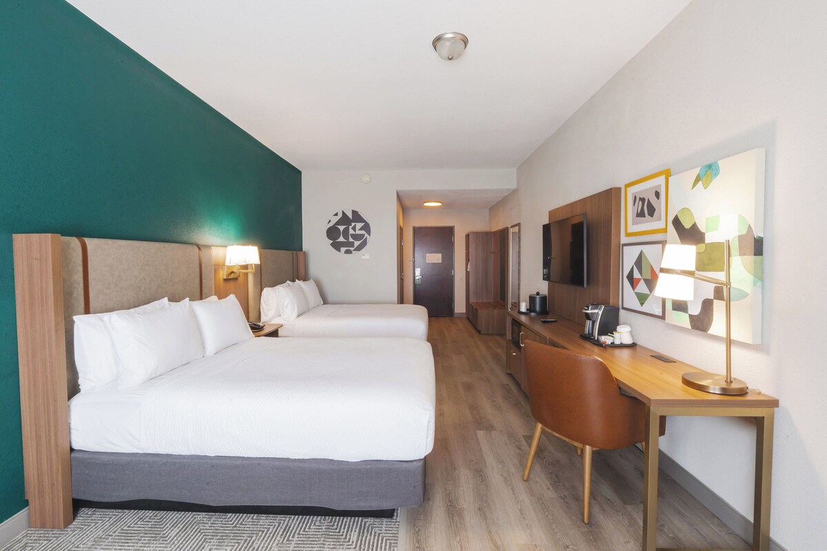 A hotel room with two beds, a desk, and a chair. There is a television on the desk and a lamp on the wall.
