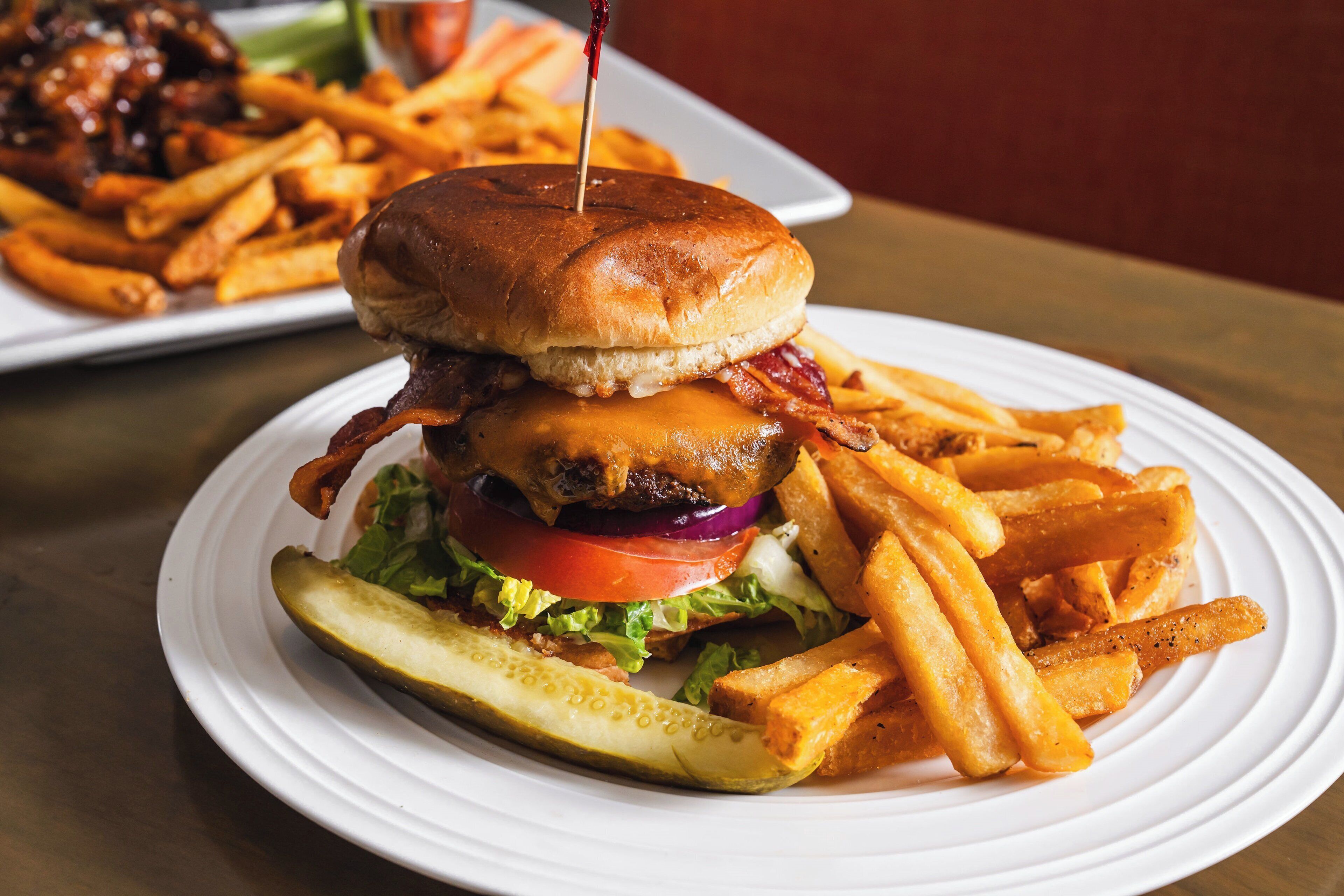 A burger with bacon, lettuce, tomato, and a pickle on a white plate, accompanied by French fries.