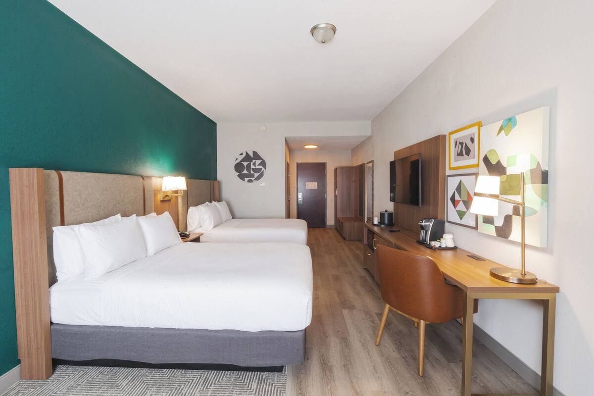 A hotel room with two beds, a desk, and a chair. There is a television on the desk and a lamp on the wall.