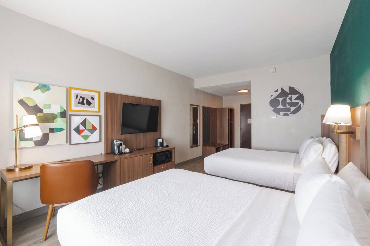 A hotel room with two beds, a desk, a TV, and artwork on the wall.