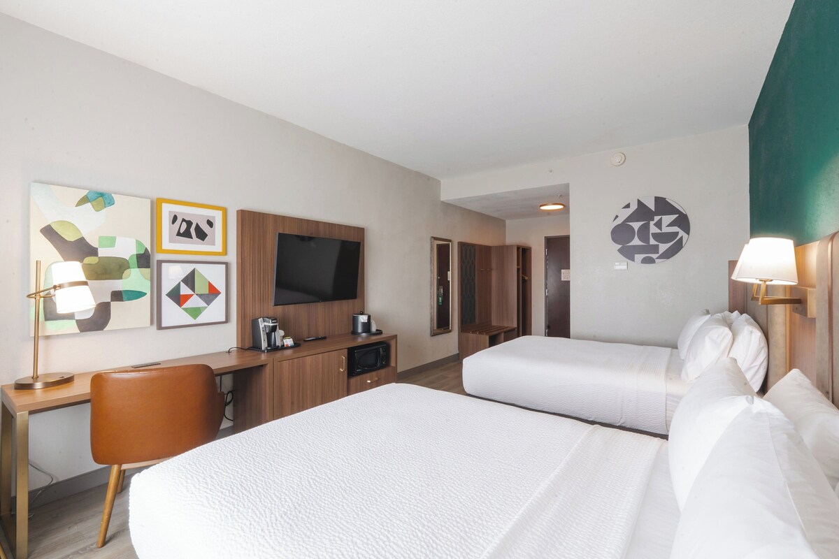A hotel room with two beds, a desk, a TV, and artwork on the wall.