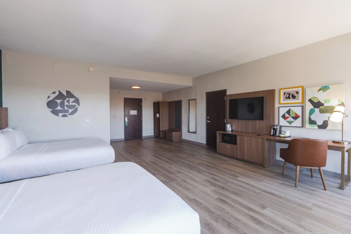 A hotel room with two beds, a kitchenette, a desk, and artwork on the walls.