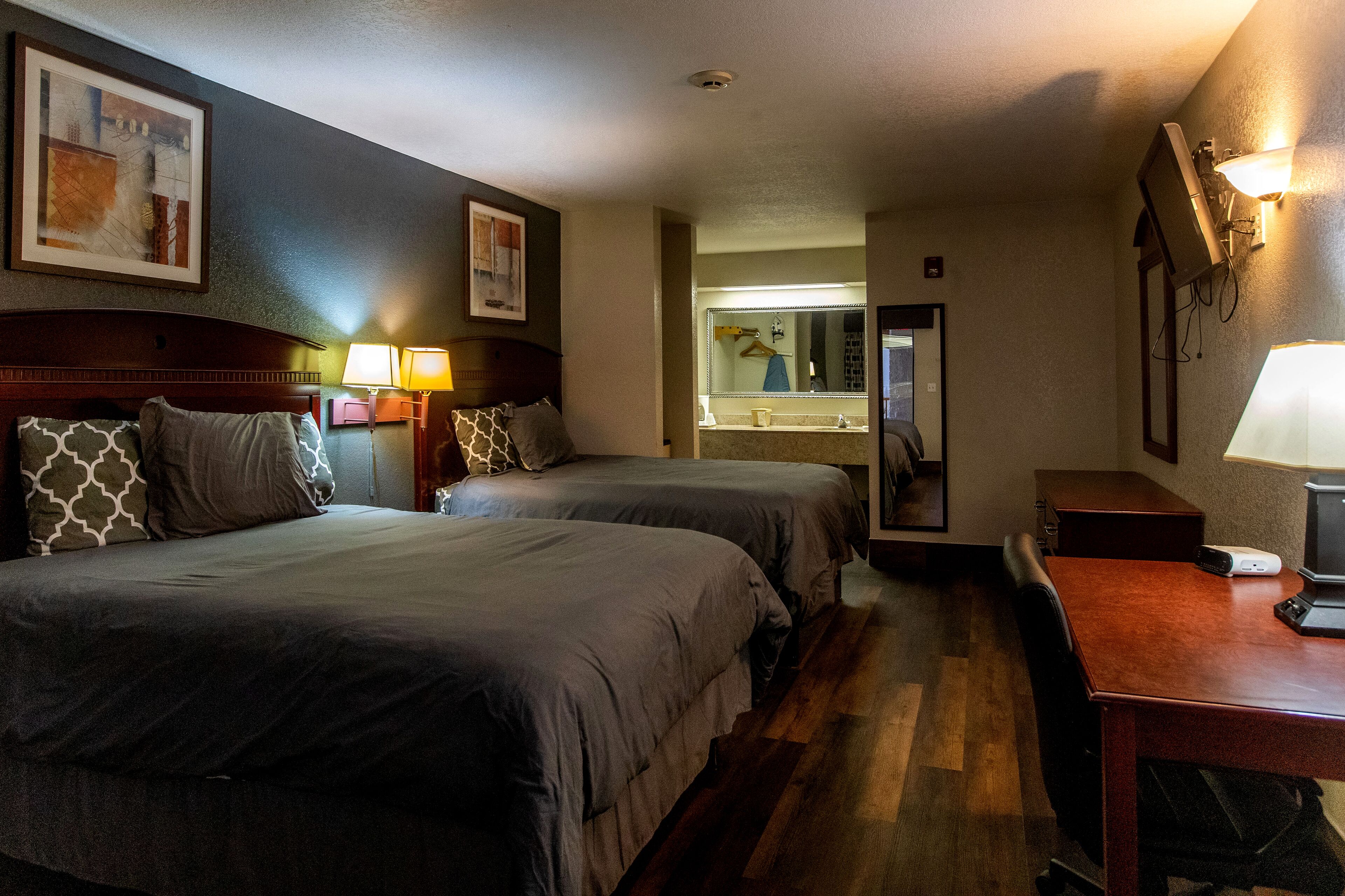 A hotel room with two beds, a desk, a mirror, and a bathroom visible through an open door.