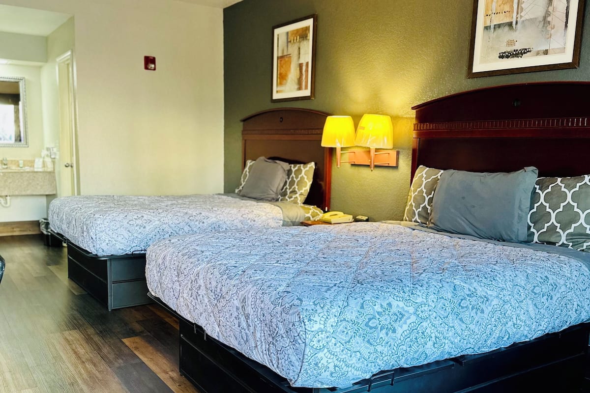 A hotel room with two beds, wooden flooring, a nightstand with lamps, and a framed picture on the wall.