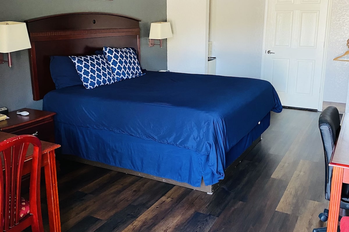 A hotel room with a large bed, wooden headboard, two bedside lamps, a desk with a chair, and a door.