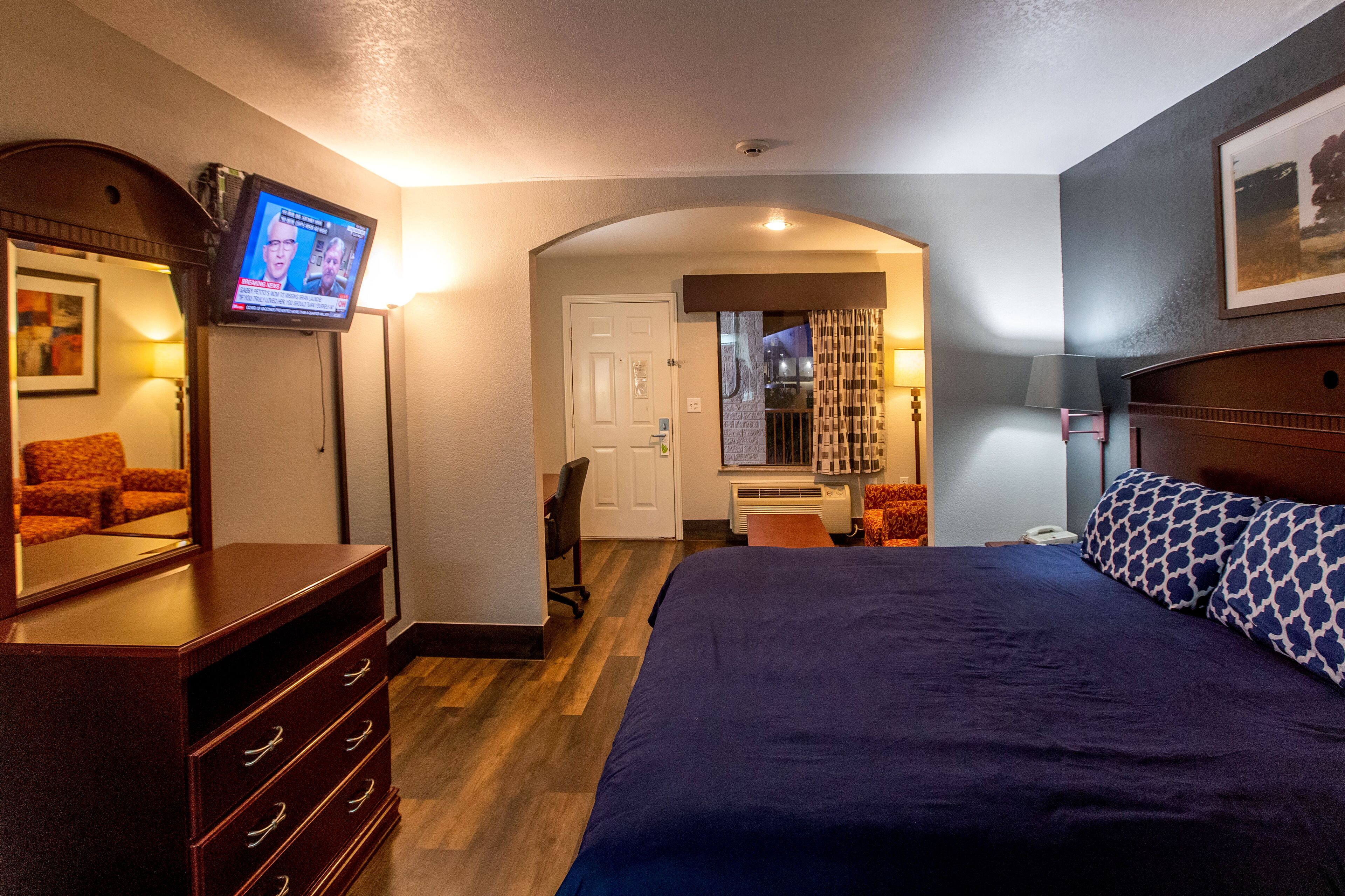 A hotel room with a large bed, a dresser, a TV mounted on the wall, a desk with a chair, and a window with curtains.