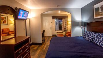 A hotel room with a large bed, a dresser, a TV mounted on the wall, a desk with a chair, and a window with curtains.